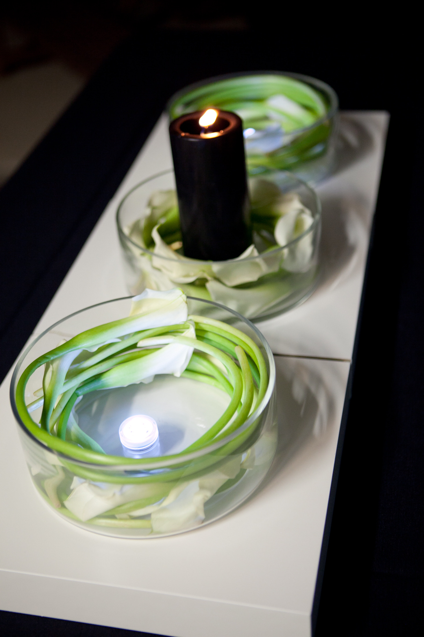 Finished centerpiece — low bowl vases with coiled green stems, white calla lily petals, submersible LED, and black pillar candle on white platform