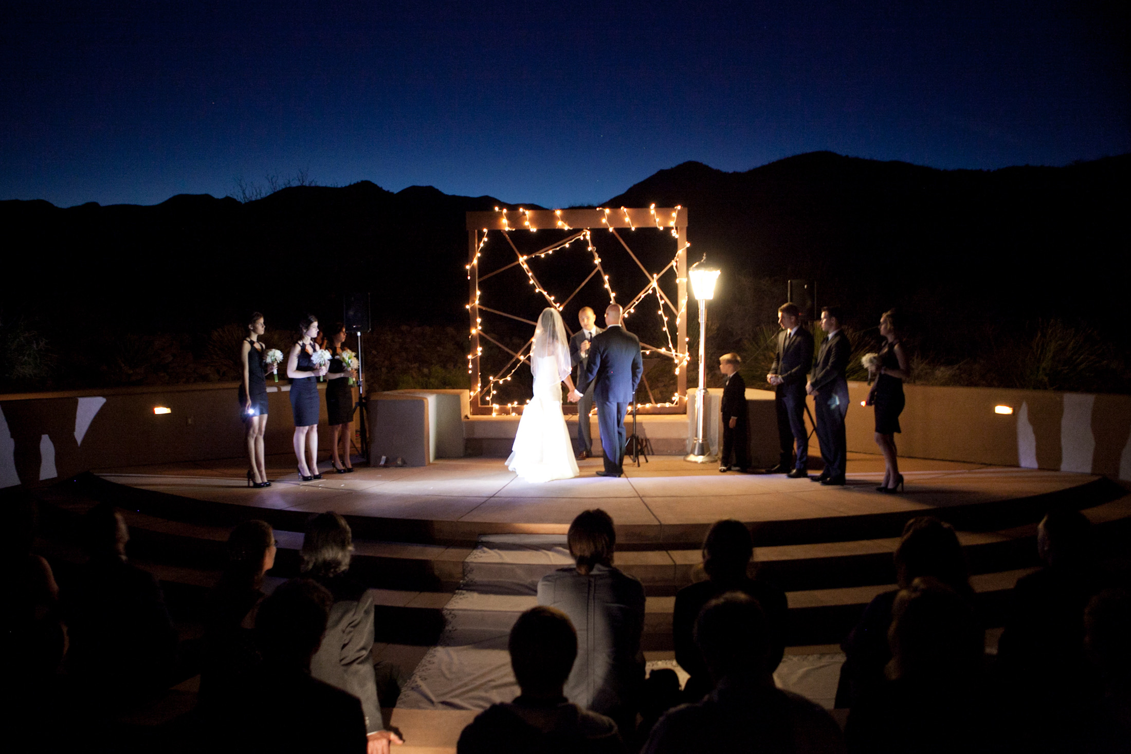 Ceremony stage — outdoor amphitheater, Arizona desert at night, showing full wedding party, audience, and custom lamppost